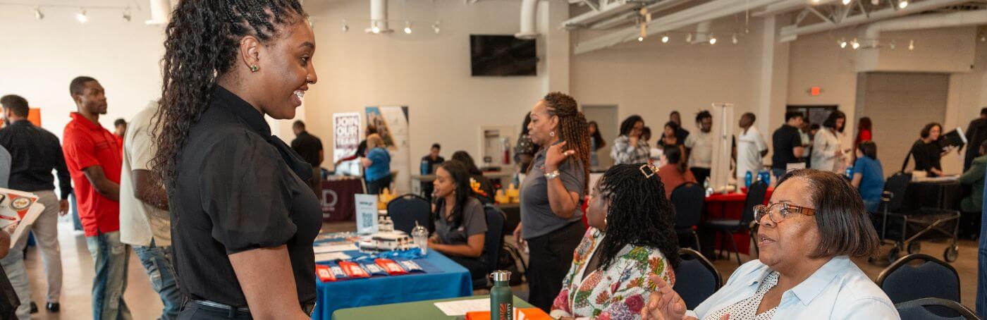 A young woman smiling while speaking with attendees at an indoor job fair or community networking event