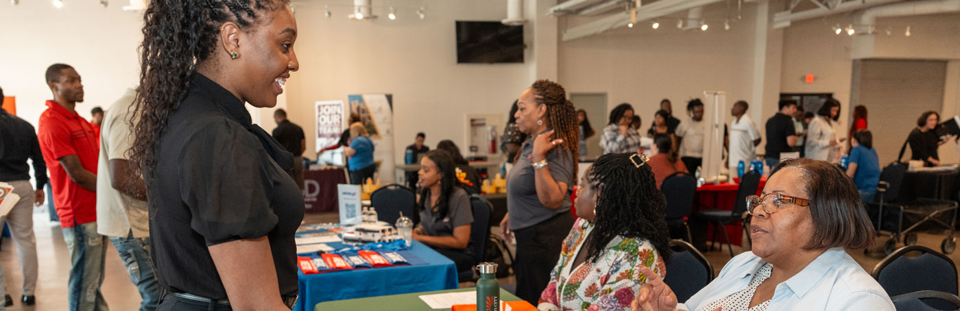 A young woman smiling while speaking with attendees at an indoor job fair or community networking event